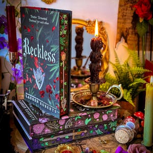 May include: A stack of books with the title "Reckless" and the author's name "Lauren Roberts" is displayed. The books have floral and botanical designs in shades of pink, green, and purple. A lit candle and decorative items surround the books, creating a mystical aesthetic.
