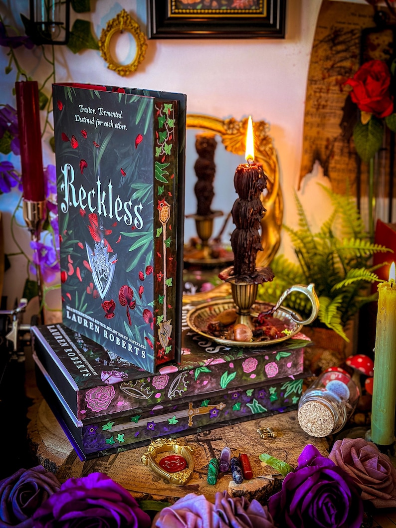 May include: A stack of books with the title "Reckless" and the author's name "Lauren Roberts" is displayed. The books have floral and botanical designs in shades of pink, green, and purple. A lit candle and decorative items surround the books, creating a mystical aesthetic.