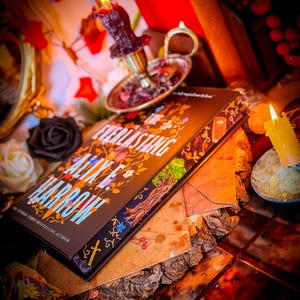 May include: A hardcover book titled "The Everlasting" by Alix E. Harrow, with a decorative cover featuring floral and tree illustrations. The book rests on a wooden surface, surrounded by candles, a black rose, and a decorative candle holder.