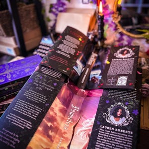 May include: Several copies of the book "Powerless" by Lauren Roberts are displayed, with open pages revealing text and cover art. The covers feature a woman with a bow and arrow, and the book is surrounded by decorative elements.