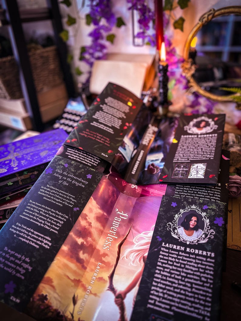 May include: Several copies of the book "Powerless" by Lauren Roberts are displayed, with open pages revealing text and cover art. The covers feature a woman with a bow and arrow, and the book is surrounded by decorative elements.