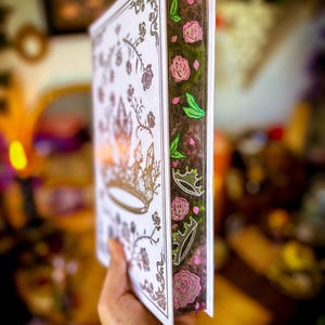May include: A decorative book with a white cover featuring a gold crown and floral design. The spine is dark brown with pink roses, green leaves, and crowns. The book is held in a hand, with a blurred background.