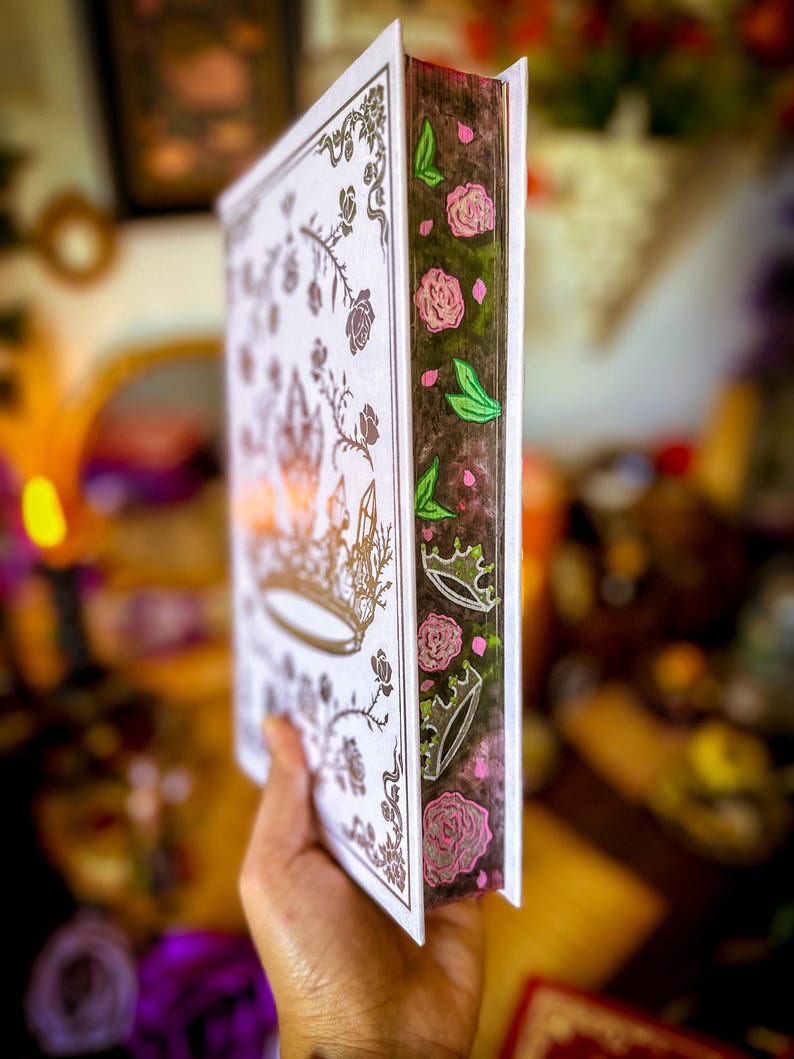 May include: A decorative book with a white cover featuring a gold crown and floral design. The spine is dark brown with pink roses, green leaves, and crowns. The book is held in a hand, with a blurred background.