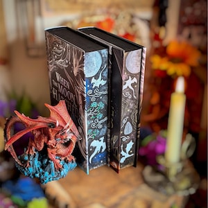 May include: Two hardback books with dark covers featuring celestial and botanical illustrations. The spine reads "When the Moon Hatched." A red dragon figurine rests on a blue crystal base in front of the books. The books are on a wooden surface.