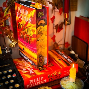 Puede incluir: Libros titulados "The Book of Dust" de Philip Pullman, con cubiertas rojas y naranjas. Un marcapáginas decorativo de gato está encima de un libro. También se ven una vela encendida y una máquina de escribir vintage.