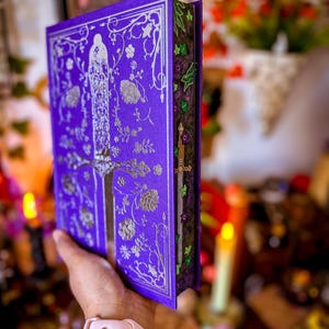 May include: A purple book with silver floral and figure designs. The book's cover features an ornate pattern with a central figure and floral accents. The edges of the pages are decorated with green and purple floral designs. A sword is visible on the side.
