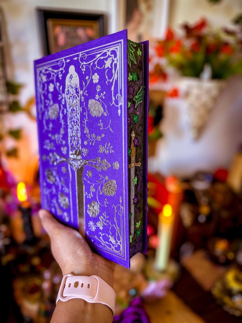 May include: A purple book with silver floral and figure designs. The book's cover features an ornate pattern with a central figure and floral accents. The edges of the pages are decorated with green and purple floral designs. A sword is visible on the side.