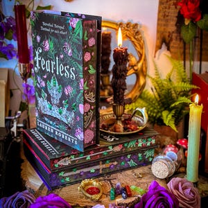 May include: A stack of books with the title "Fearless" by Lauren Roberts, featuring a floral and crown design. The books are on a wooden surface, surrounded by purple and pink roses, candles, and decorative items.