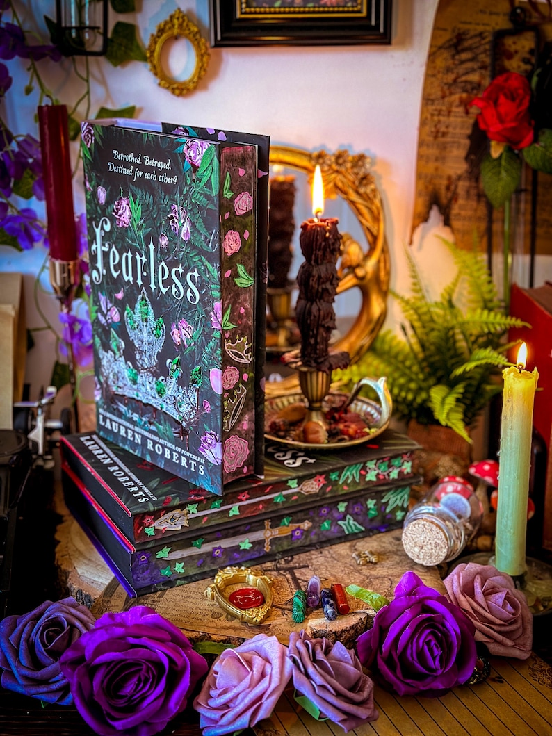 May include: A stack of books with the title "Fearless" by Lauren Roberts, featuring a floral and crown design. The books are on a wooden surface, surrounded by purple and pink roses, candles, and decorative items.
