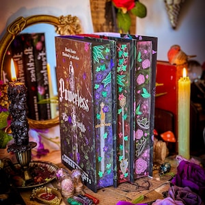 Può includere: Una pila di libri fantasy con copertine decorative. I dorsi dei libri presentano motivi floreali e di spade in viola, verde e rosa. Il libro intitolato "Powerless" è visibile, insieme ad altri titoli. La scena comprende candele, rose e oggetti decorativi.
