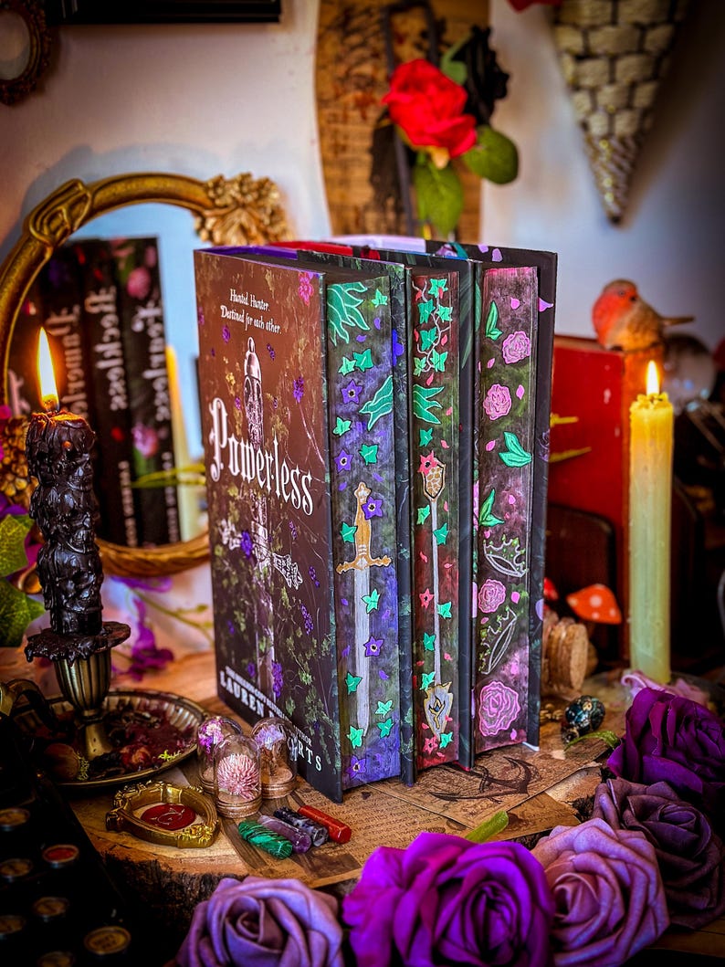 May include: A stack of fantasy books with decorative covers. The book spines feature floral and sword designs in purple, green, and pink. The book titled "Powerless" is visible, along with other titles. The scene includes candles, roses, and decorative items.