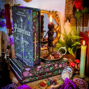 May include: A stack of decorative book boxes and a hardcover book titled "Powerless" by Lauren Roberts. The boxes and book feature floral and botanical designs in shades of purple, green, and black. Candles and roses surround the books.