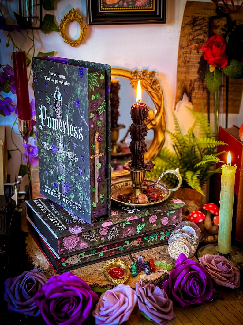 May include: A stack of decorative book boxes and a hardcover book titled "Powerless" by Lauren Roberts. The boxes and book feature floral and botanical designs in shades of purple, green, and black. Candles and roses surround the books.