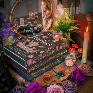May include: A stack of books with the title "Fearless" on the cover, surrounded by decorative elements. The books have a black cover with floral and botanical designs. Purple and pink roses are arranged in the foreground, with candles, a mirror, and other decorative items.
