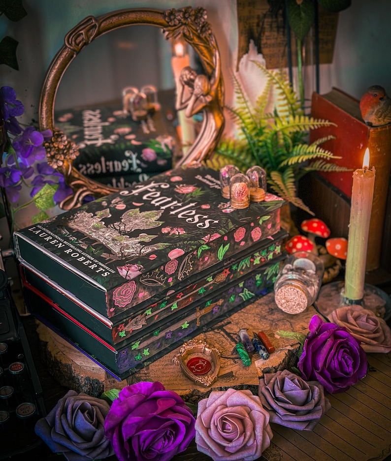 May include: A stack of books with the title "Fearless" on the cover, surrounded by decorative elements. The books have a black cover with floral and botanical designs. Purple and pink roses are arranged in the foreground, with candles, a mirror, and other decorative items.