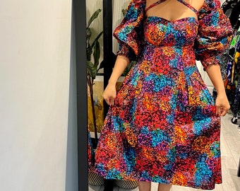 May include: A woman wearing a colorful, patterned midi dress with puffed sleeves and a criss-cross halter neckline. She is wearing silver heels and standing in front of a mirror.