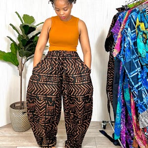 May include: A woman wearing a mustard-colored tank top and black patterned pants with a brown geometric design. The pants have a loose, baggy fit. A rack of colorful clothing hangs in the background.