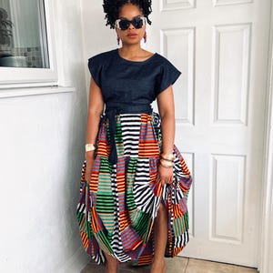 May include: A dark blue short-sleeved top with a matching tie-waist belt paired with a voluminous, floor-length skirt featuring a vibrant, multi-colored patchwork pattern of horizontal stripes in red, yellow, green, black, and white.