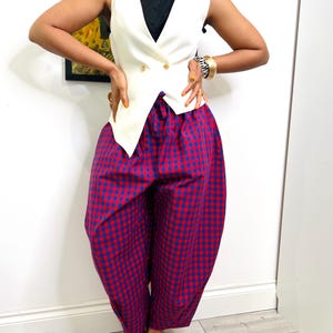 May include: A cream waistcoat with gold buttons worn over a black collared shirt. The outfit includes red and blue checked trousers and zebra-print heels. A striking fashion ensemble.