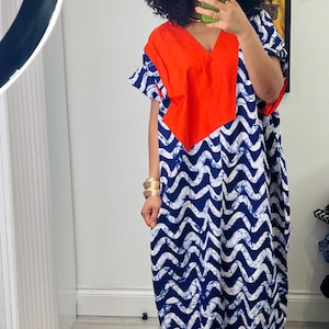 May include: A long, loose-fitting dress featuring a V-neck and a striking colour-block design. The top section is a bold orange, complemented by a blue and white wave pattern. The dress has short sleeves and is designed for comfort, ideal for warmer climates.