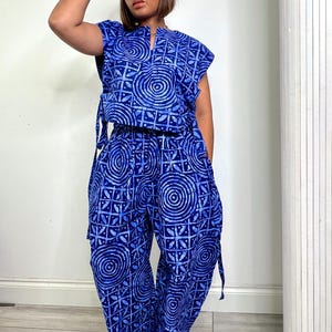 Puede incluir: Un conjunto de dos piezas compuesto por una blusa de manga corta y pantalones de pierna ancha, ambos con un vibrante estampado estilo batik en azul y blanco con círculos concéntricos y formas geométricas. La blusa tiene cuello con abertura.