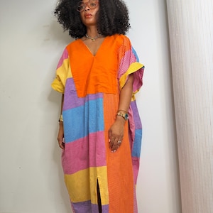 May include: A loose-fitting, ankle-length kaftan dress featuring a vibrant patchwork design of orange, yellow, blue, and pink panels. The garment has a V-neckline, short sleeves, and a central front slit, crafted from a mix of solid and fine-checkered fabrics.