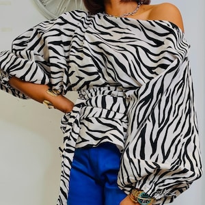 May include: A long-sleeved blouse featuring a black and white zebra print pattern, an off-the-shoulder neckline, and a wrap-around waist tie, paired with solid blue trousers. The blouse has voluminous sleeves with gathered cuffs.