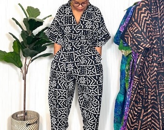 May include: A black and white patterned jumpsuit with a wrap-style top and wide-leg pants. The jumpsuit features a geometric design and is worn with platform sandals. Other patterned garments hang on a rack in the background.
