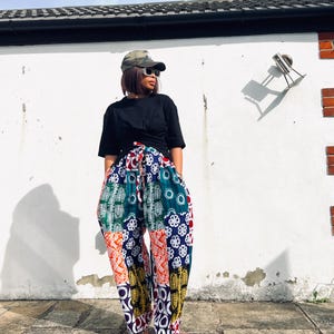 May include: A black short-sleeved t-shirt paired with loose-fitting, high-waisted trousers featuring a vibrant patchwork pattern of blue, orange, green, and yellow geometric and floral motifs, complete with a drawstring waist and a camouflage baseball cap.
