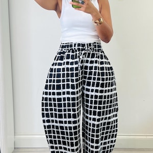 Puede incluir: Una camiseta sin mangas blanca combinada con pantalones anchos y holgados con un patrón de cuadrícula en blanco y negro. Los pantalones tienen una cintura elástica con cordón. El conjunto se complementa con una gorra de béisbol negra.