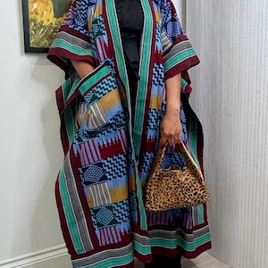 May include: A long, open-front kimono-style robe featuring a vibrant, multi-colored geometric pattern in shades of burgundy, blue, yellow, and green. The garment includes wide sleeves, a solid-colored trim, and two large front patch pockets.