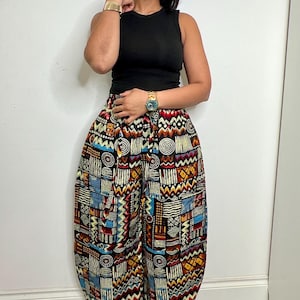 Puede incluir: Una persona lleva una camiseta sin mangas negra y pantalones anchos estampados. Los pantalones presentan un diseño geométrico colorido en rojo, azul, amarillo y negro. La persona también lleva una gorra verde, gafas y un reloj dorado.