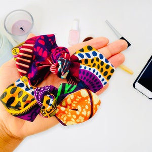 以下が含まれることがあります： 鮮やかなアフリカンプリント柄の生地で作られたカラフルなヘアゴム。ヘアゴムは結び目があり、紫、黄、オレンジ、緑の色が混ざり合っています。