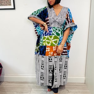 May include: A vibrant patchwork caftan dress with a V-neck, showcasing a variety of patterns and colours such as blue, green, orange, and white. The dress has wide sleeves and a long skirt. A green cap with white text is worn.