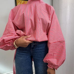 May include: A high-neck blouse featuring a red and white vertical striped pattern, long voluminous sleeves with gathered cuffs, and a loose-fitting bodice paired with dark blue denim jeans. The blouse includes side ties and a structured collar.