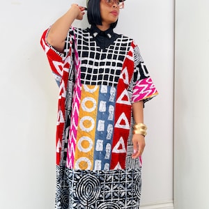 May include: A vibrant, patterned dress with a black collar and short sleeves. The dress showcases geometric shapes, including squares, triangles, and circles, in red, white, black, yellow, and blue. The outfit is completed with a green baseball cap and gold bangles.