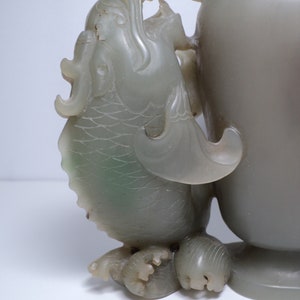 May include: A jade vase with a dragon handle. The vase is a pale green color and has a smooth, polished finish. The dragon is carved in detail and has a fierce expression.