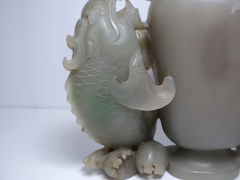 May include: A jade vase with a dragon handle. The vase is a pale green color and has a smooth, polished finish. The dragon is carved in detail and has a fierce expression.