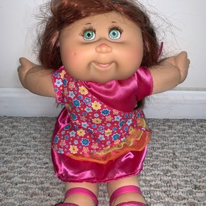 Pode incluir: Uma boneca Cabbage Patch Kid com cabelo castanho, olhos verdes e um vestido rosa com padrão floral. A boneca está usando sapatos rosa.