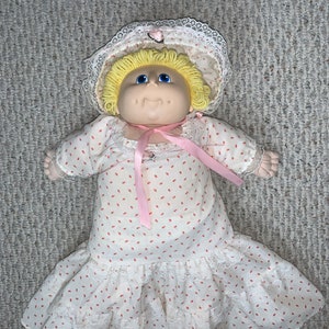 Puede incluir: Una muñeca Cabbage Patch Kid con un vestido blanco con lunares rosas y un borde de encaje blanco. La muñeca tiene el pelo rubio y los ojos azules y lleva una capota de encaje blanco con un lazo rosa.