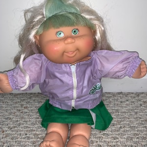 Peut inclure: Une poupée Cabbage Patch Kid avec des cheveux blonds et des yeux verts portant une veste violette avec une fermeture éclair blanche et une jupe verte. La poupée est assise sur un tapis vert.