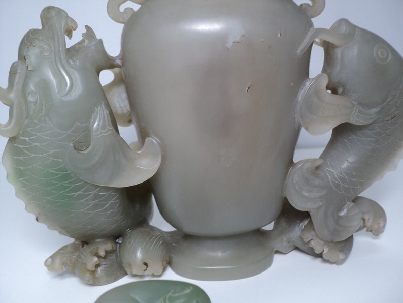 May include: A pale green jade vase with two fish handles. The vase is carved with intricate details and has a smooth, polished finish.