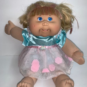 Op de afbeelding: Een Cabbage Patch Kid pop die een lichtblauwe satijnen jurk draagt met een roze tule overlay en roze bloemaccenten. De pop heeft blond haar met roze highlights en blauwe ogen.