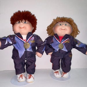 May include: Two Cabbage Patch Kids dolls dressed in blue and purple tracksuits with red, white, and blue accents. Both dolls are wearing gold medals around their necks. The dolls are standing on white bases.