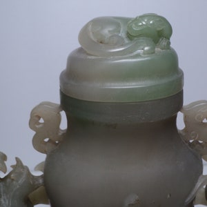 May include: A jade green vase with a lid featuring a carved animal. The vase has two handles on either side, each with a carved animal design. The vase is made of a smooth, polished jade.