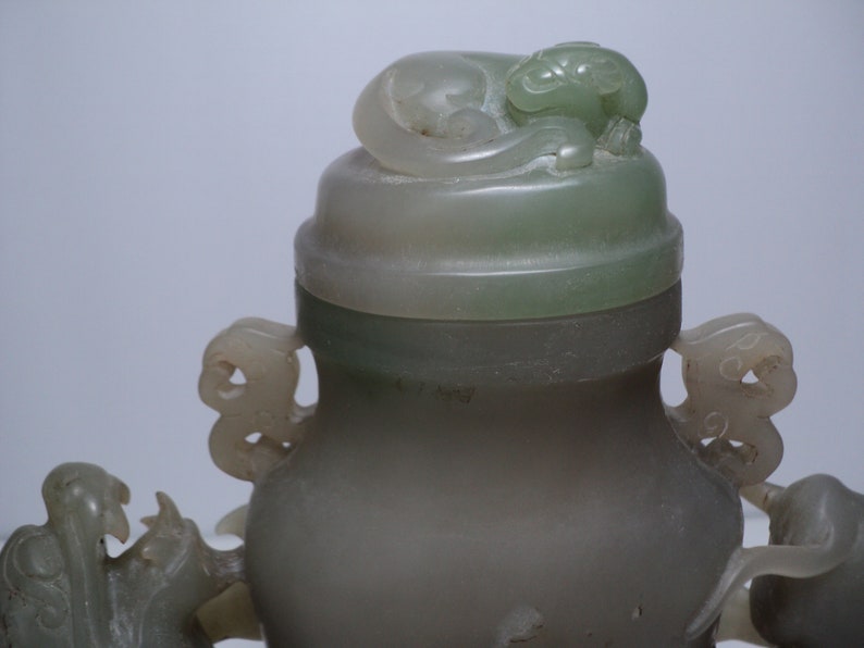 May include: A jade green vase with a lid featuring a carved animal. The vase has two handles on either side, each with a carved animal design. The vase is made of a smooth, polished jade.