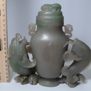 May include: A jade green vase with a lid and two fish handles. The vase is carved with intricate details and has a smooth, polished finish.