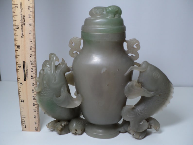 May include: A jade green vase with a lid and two fish handles. The vase is carved with intricate details and has a smooth, polished finish.