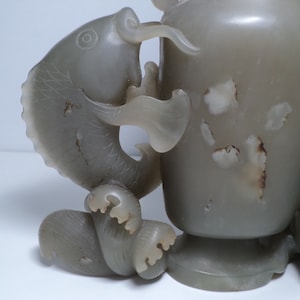 May include: A jade vase with a fish design. The vase is a light gray color and has a smooth, polished finish. The fish is carved in detail and has a realistic appearance.