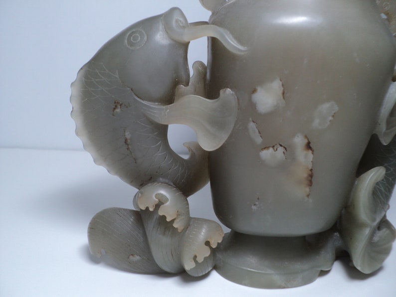 May include: A jade vase with a fish design. The vase is a light gray color and has a smooth, polished finish. The fish is carved in detail and has a realistic appearance.